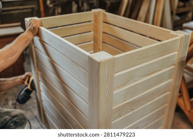Partial view of a craftsperson sanding a large unfinished slatted pine planter or storage box in a woodworking workshop, with visible grain and sturdy corner posts