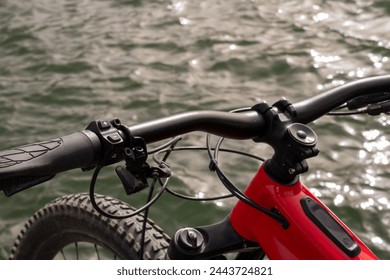 Partial view of an e-bike (handlebar, red frame, tires) by the water - Powered by Shutterstock