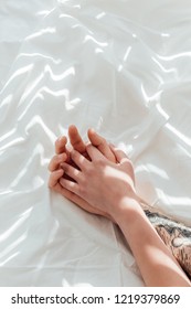 Partial View Of Loving Couple Holding Hands While Lying On White Bed Sheet
