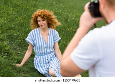partial view of man taking picture of smiling redhead woman on grassy meadow