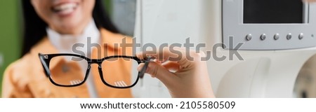 partial view of oculist holding eyeglasses near smiling woman and blurred vision screener, banner