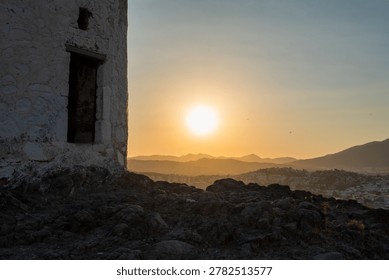 Partial view of an old stone windmill ruin on a rocky hill overlooking a hazy coastal town and mountains during a golden sunset