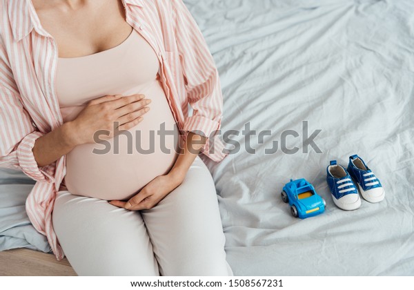 partial view of pregnant woman with\
toy car and baby shoes sitting on bed and touching\
belly