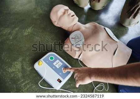 partial view of professional medical instructor operating defibrillator on CPR manikin near young participants of first aid seminar, high angle view, health care and life-saving techniques concept