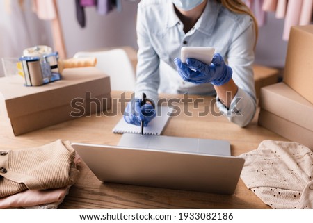 partial view of proprietor in latex gloves working with smartphone and laptop in showroom, blurred background