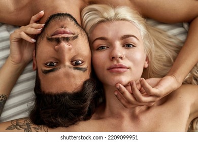 Partial View Of Serious Young Beautiful Multiethnic Couple Lying And Looking At Camera On Bed At Home. Caucasian Girl And Middle Eastern Man Touching Each Other. Domestic Lifestyle. Closeness