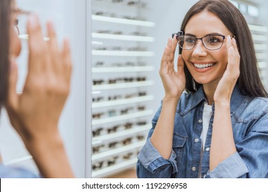 partial view of smiling woman choosing eyeglasses and looking at mirror in optica 