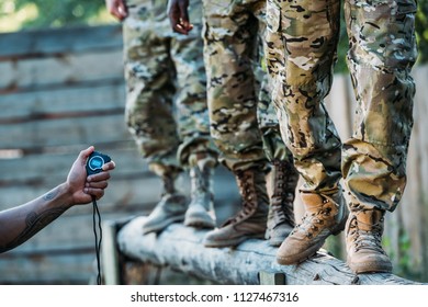 partial view of tactical instructor with stop watch examining multiracial soldiers during obstacle run on range