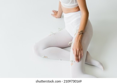 Partial View Of Tattooed Woman In White Tights With Tiny Flowers Sitting On Light Grey