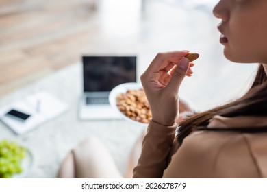 partial view of woman eating almonds near blurred laptop and smartphone on floor