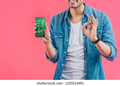 partial view of young man doing ok sign and showing smartphone with medical application isolated on pink