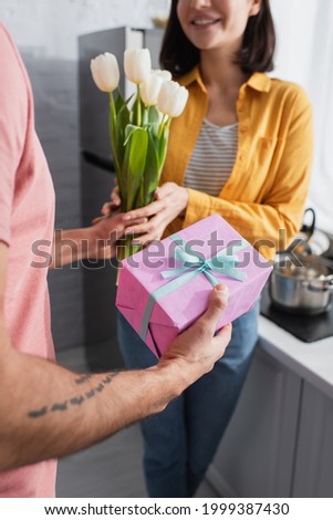 partial view of young man presenting bouquet of flowers and gift box to girlfriend in kitchen