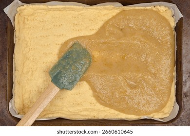 A partially baked pie crust in a rectangular baking pan, with cooked apple filling being spread onto it using a spatula