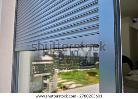 Partially closed gray roller shutter covers window on modern home exterior. Residential privacy, sun shade and security concept.