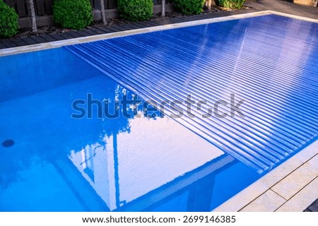 A partially closed slatted pool shutter. The water's surface reflects the building's façade and tree canopies. The shot conveys the cleanliness and well-maintained nature of the private courtyard.