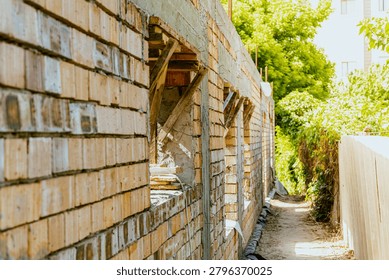 Partially constructed brick wall with open window frames, surrounded by lush greenery and a clear sky, showcasing a construction site in progress