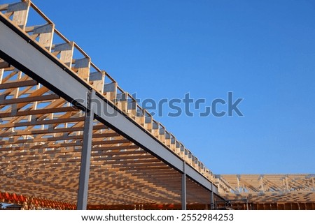 A partially constructed building featuring a combination of steel beams and wooden trusses, showcasing modern architectural design and construction techniques under a clear blue sky.