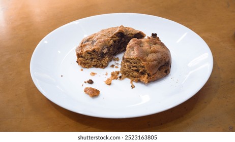 A partially eaten cookie on a white plate is placed on a wooden surface. The cookie appears golden brown with visible chunks, suggesting it's a chocolate chip or nut-filled variety. Crumbs are scatter - Powered by Shutterstock