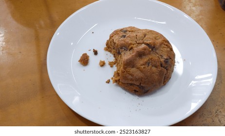 A partially eaten cookie on a white plate is placed on a wooden surface. The cookie appears golden brown with visible chunks, suggesting it's a chocolate chip or nut-filled variety. Crumbs are scatter - Powered by Shutterstock