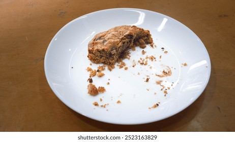 A partially eaten cookie on a white plate is placed on a wooden surface. The cookie appears golden brown with visible chunks, suggesting it's a chocolate chip or nut-filled variety. Crumbs are scatter - Powered by Shutterstock