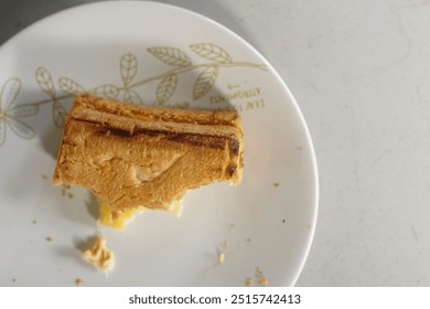 Partially eaten grilled cheese sandwich on white plate with golden leaf design. Toasted bread reveals melted cheese inside. Crumbs scattered on plate. Comfort food closeup on light background. - Powered by Shutterstock