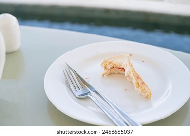 A partially eaten sandwich with jam on a white plate, accompanied by a fork and knife, placed on a light green table near a pool. - Powered by Shutterstock