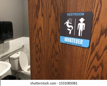 Partially open restroom door and sign with gender inclusive language and symbols stating WHATEVER JUST WASH YOUR HANDS. Toilet, sink, grab bar and towel dispenser are seen inside.
