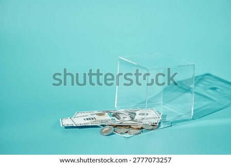 A partially open transparent cube with coins and dollar bills half inside, half spilled out on turquoise background. Symbol of insecure storage, mixed currency risk, and lost savings. Copy space for text.