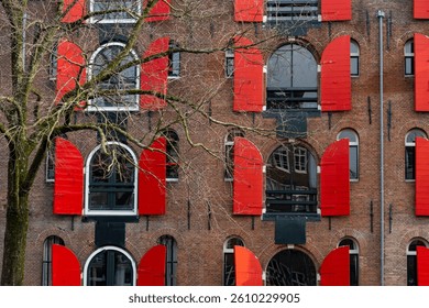 Partially opened red window shutters create a vibrant contrast against the brown brick facade of a typical amsterdam building