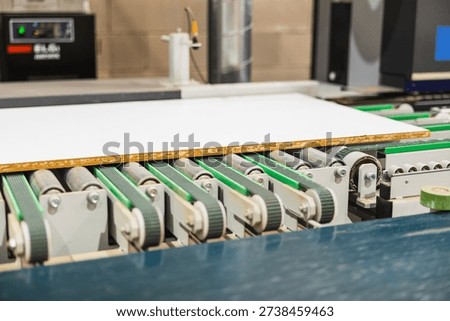 Particle board panel with a white surface moving on a conveyor system with green belts and metal rollers in a production plant