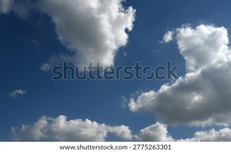 Partly clouded sky with large cloud formations and open blue sky between them, showing a gradient from dark blue at the top to lighter blue at the bottom. Sunlight shines from the upper right corner, casting a shadow under the left foremost cloud.