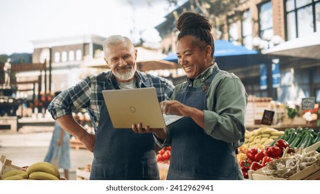 Partners Working at a Farmers Market. Middle Aged Couple Selling Ecological Fruits and Vegetables From a Food Stand. Street Vendors Using Laptop Computer and Discussing Steps for Business Development - Powered by Shutterstock