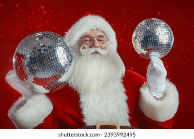 Party for Christmas and New Years. Cheerful Santa Claus holds two shiny disco balls in his hands, rejoices and says ho-ho-ho. Red holiday background with copy space. - Powered by Shutterstock