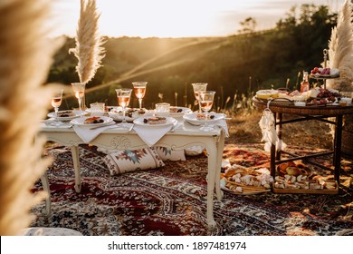 Party Festival Table Outdoor für Feiertage Feiertage. Trinken Sie Gläser und süßen Dessert auf Holzsteg, Kissen, Teppich und trockene Pflanzendekorationen. Fest und Treffen, Sonnenuntergang auf Hintergrund