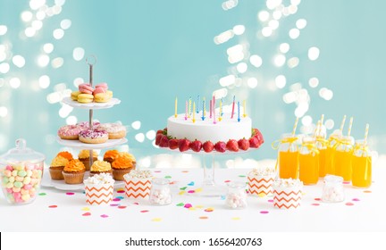 Party And Festive Concept - Birthday Cake With Candles And Strawberries, Drinks And Food On Table Over Lights On Blue Background