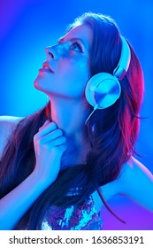 Party and holiday style. Pretty girl with shiny make-up and shiny dress listening to music in headphones and dancing in blue and pink light.