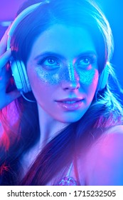 Party and holiday style. Pretty girl with shiny make-up and shiny dress listening to music in headphones and dancing in blue and pink light.