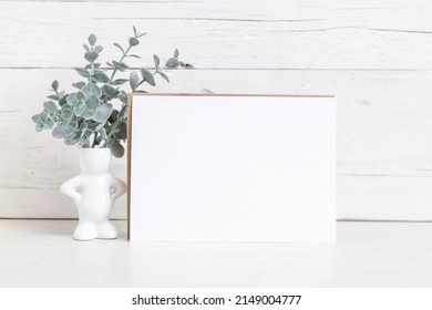 Party Invitation Mockup On A Light Background With Flowers.  Horizontal White Card 5x7 On The Table Against A Light Wall.