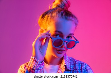 Party. Pretty young girl, student in cool eyeglasses wearing plaid shirt isolated on magenta color background in neon light. Concept of beauty, art, fashion, youth, sales and ad, education, studying. - Shutterstock ID 2181370941