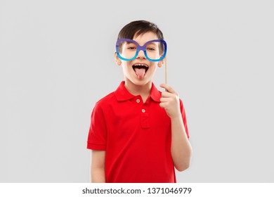 Party Props, Photo Booth And Childhood Concept - Happy Little Boy In Red Polo T-shirt With Big Paper Glasses Showing Tongue Over Grey Background