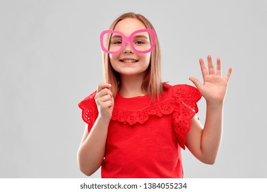 Party Props, Photo Booth And Childhood Concept - Smiling Girl In Red Shirt With Big Paper Glasses Waving Hand Over Grey Background
