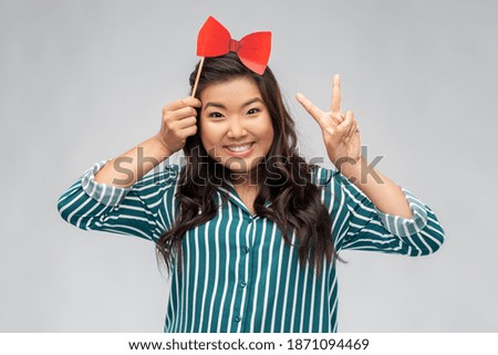 party props, photo booth and people concept - happy asian young woman with red paper bow showing peace hand sign over grey background