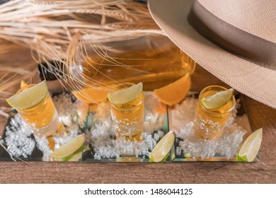 Party At The Ranch, A White Hat On Top Of Three Shots Of Whiskey, Vodka And Tequila With Slices Of Ripe Orange, Lime Green With Salt On The Wooden Table. Straw Bunches Covering A Bottle Of Vodka. 