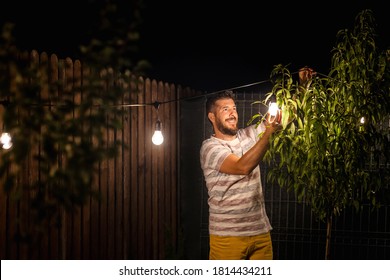 Party Time In Backyard With Happy Man Hanging String Lights In Trees – Weekend Night Mood With Smiling Millennial Guy Arranging The Light Garland For Outdoor Dinner Party In Garden