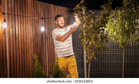 Party Time In Backyard With Happy Millennial Man Hanging String Lights In Trees – Weekend Summer Night Mood With Smiling Guy Arranging The Light Garland For Outdoor Dinner Party In Home Garden