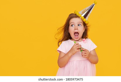 Party time. A joyful little girl in a festive cap and elegant dress celebrates her birthday. Blowing a whistle on a yellow background