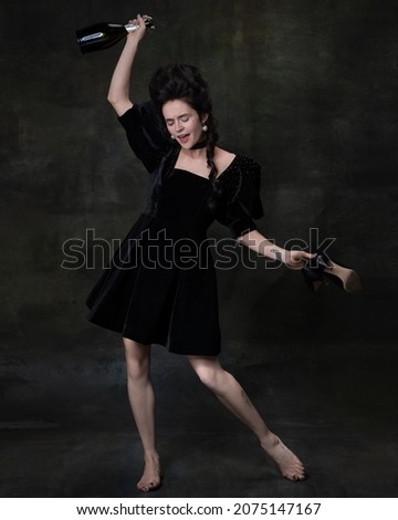 Party time, tipsiness. Full-length portrait of young beautiful happy woman in black attire drinking wine and laughing isolated on dark vintage background. Concept of comparison of eras, modernity Foto stock © 