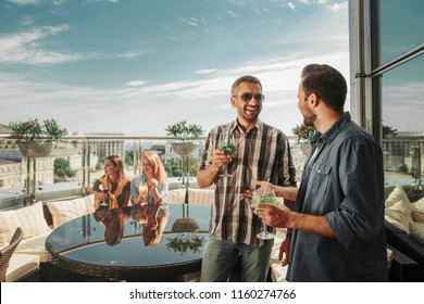 Party under the sky. Smiling friends enjoying cocktails at rooftop bar. Gentlemen talking while lovely women sitting on the couch with cushions - Powered by Shutterstock