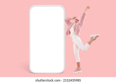 Partying girl on pink background in fake fur bomber and stylish shoes feeling light as feather dancing next to vertical gigantic phone template with blank copy space for your promotional content