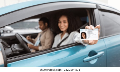 Passed Exam. Happy middle eastern woman sitting in car showing driver license from window, celebrating driving school finish, inside of vehicle with instructor man on passenger seat. Focus on card - Shutterstock ID 2322194671